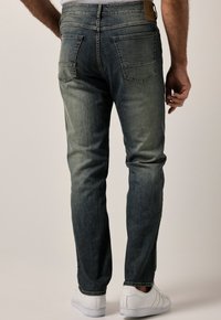 Vaqueros de mezclilla azul desgastados con un corte ajustado, que cuentan con bolsillos traseros y un parche de cuero con marca en la cintura. Zapatillas blancas completan el look.