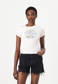 Camiseta de manga corta blanca con relieve que presenta el texto "Tommy Jeans New York" en azul, rojo y negro, combinada con pantalones cortos de mezclilla negros con bordes deshilachados.