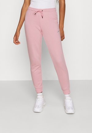 Vrouw draagt lichtroze Nike joggingbroek, witte sokken, witte sneakers en een wit shirt, staand tegen een effen lichte achtergrond.