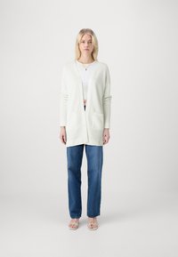 Gara, gaiši zaļa kardigāna, ar faktūras adījuma rakstu, atvērta priekšpuse un divas priekšējās kabatas, saskaņota ar zilo denim auduma biksēm.