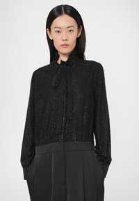 Blouse noire texturée avec manches longues, col à volants et lien au cou. Partie inférieure lisse avec plis et poches latérales.