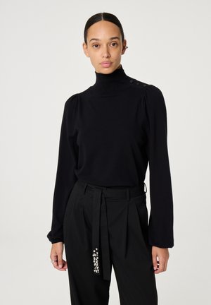 Zwarte coltrui met lange mouwen, zachte textuur en knopen op de schouders. G gedragen met hoge taille zwarte broek met versieringen.