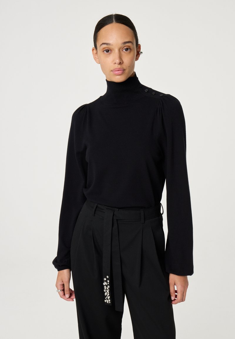 Schwarzer Rollkragenpullover mit langen Ärmeln, weicher Textur und Knopfdetails an den Schultern. Getragen mit hochgeschnittenen schwarzen Hosen mit Verzierungen.