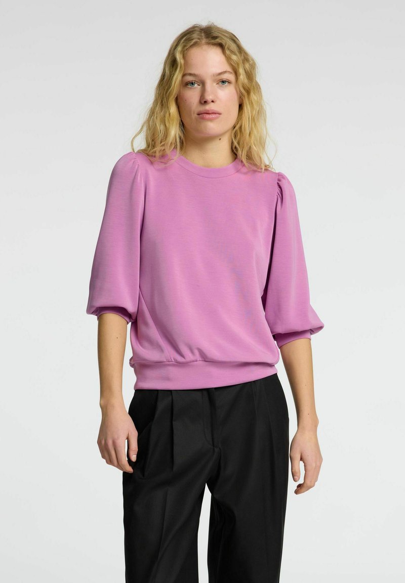 Pullover rosa con maniche corte a sbuffo e collo rotondo; presenta una vita aderente. Abbinato a pantaloni neri a vita alta.