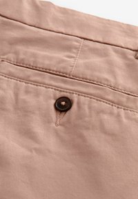 Tejido de color rosa claro con una textura suave, que cuenta con un bolsillo trasero y un botón marrón oscuro con detalle de costura.