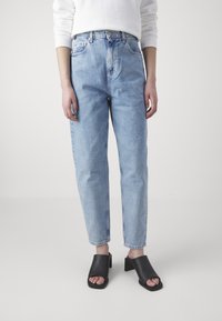 Persona vestida con jeans de tiro alto azul claro, blusa blanca de manga larga y sandalias de tacón abierto negras, de pie sobre un fondo liso.