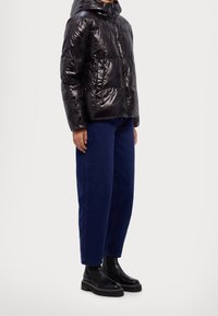 Veste matelassée noire à finition brillante, fermeture éclair à l'avant, et design rembourré. Portée avec un jean large bleu foncé et des bottines noires.