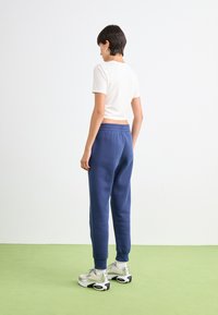 Kvinne som står på grønt gulv, iført hvit crop top, blå joggebukser, hvite sokker, sølvfargede sneakers og hoopsøredobber, vendt bort fra kamera.