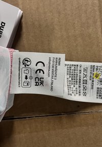 Etykieta z tkaniny, symbolem pielęgnacji i tekstem. Głównie biała z różowymi akcentami. Zawiera nazwę marki, adres oraz różne ikony zgodności.