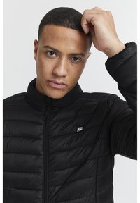 Black puffer jacket with quilted design, high collar, and front zip. Features a small logo on the chest and smooth texture.