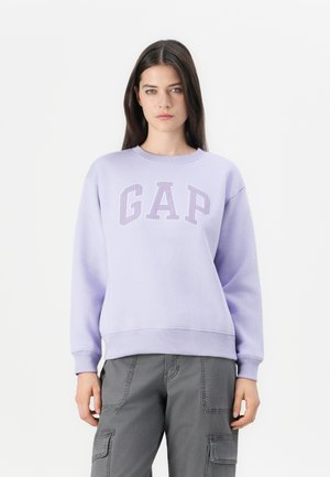 Femme aux longs cheveux bruns, portant un sweat-shirt lavande de GAP et un pantalon cargo gris, se tenant devant un fond blanc uni.