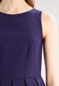 Marinin sininen hihaton mekko, jossa on pyöreä pääntie, sileä pinta ja laskostettu hameosa, joka korostaa istuvaa yläosaa.