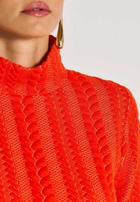 Rotes, strukturiertes Oberteil mit hohem Kragen, das über kunstvoll gewebte Muster und kontrastierendes Nähen verfügt. Eine goldene Ohrringe sorgt für einen eleganten Akzent.