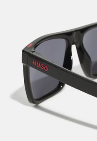Occhiali da sole neri con lenti scure, caratterizzati da un audace marchio "HUGO" in rosso sul lato. Il design ha una forma geometrica e angolare.