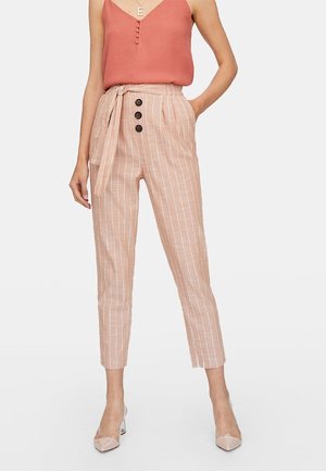 Vrouw draagt beige broek met hoge taille en dunne strepen, drie knopen en een gestrikte riem, gecombineerd met een mouwloos roestkleurig topje en nudekleurige hakken.