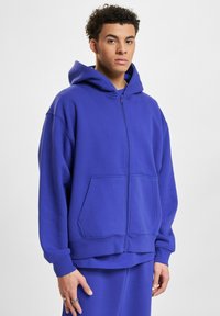 Zip-up hoodie u vibrantnoj ljubičastoj boji, izrađena od mekanog materijala. Sadrži veliku kapuljaču, prednje džepove te rebraste manžete i donji rub.