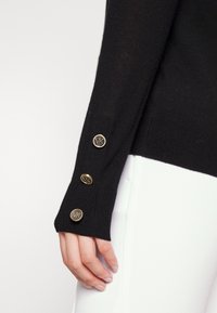 Main portant une manche noire avec trois boutons dorés embossés du logo MK près du poignet, assortie à un pantalon blanc.