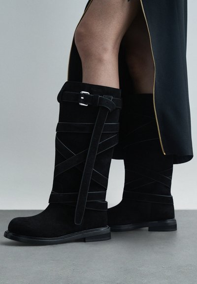Czarne zamszowe kozaki do kolan z wieloma paskami i srebrnymi klamrami, noszone z czarną spódnicą z złotym suwakiem.