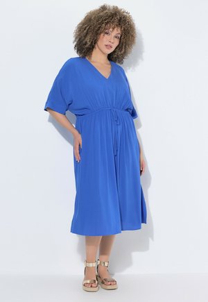 Kvinde med krøllet hår iført en kongeblå midi-kjole med korte ærmer og guld platform sandaletter med ankelrem mod en hvid baggrund.