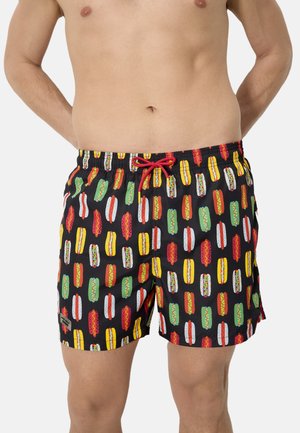 Hombre con pantalones cortos de baño negros con patrones coloridos de perritos calientes y un cordón rojo, de pie con las manos detrás de la espalda.