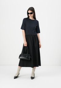Marine ribbet strikket kort topp, svart midi skjørt, svart håndveske med korte håndtak, beige sokker, svarte spisse hæler. Enkelt hvitt bakgrunn.