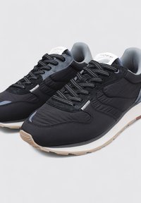 Zapatillas negras con una mezcla de materiales lisos y texturizados, con un diseño de cordones, un collar acolchado y una suela de goma de color claro.