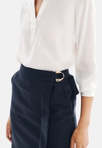 Blusa de seda blanca con escote en V, combinada con una falda oscura azul marino que presenta un detalle de hebilla circular dorada y acentos plisados.