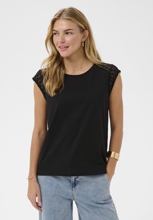 Kvinde med blondt hår iført en sort ærmeløs top med blonder på skuldrene og lyseblå jeans, smilende og kiggende til siden.