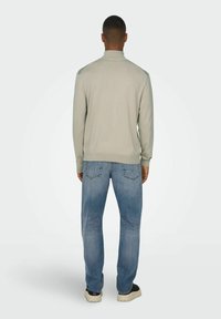 Hellgrauer Pullover mit Kragen, enganliegenden Bündchen und strukturierter Oberfläche, kombiniert mit blauen Denim-Jeans mit subtilen Ausbleichungen.