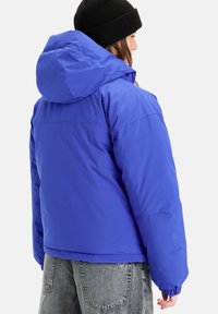 Blauer Puffermantel mit Kapuze, der eine glatte, glänzende Textur und elastische Bündchen aufweist. Über grauen Jeans getragen, zeigt er einen lässigen Schnitt.