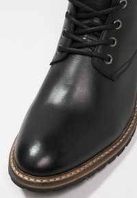 Musta nahkakengät, joissa on sileä pinta, pyöreä kärki ja kestävä pohja. Ominaisuuksiin kuuluvat metalliset silmukat ja mustat kengännauhat. Kontrastivärinen ommel kengän pohjan ympärillä.