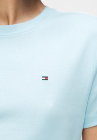 Κοντινό πλάνο ενός ατόμου που φοράει γαλάζιο T-shirt με ένα μικρό κεντημένο ορθογώνιο λογότυπο σε μπλε σκούρο, λευκό και κόκκινο στο στήθος.