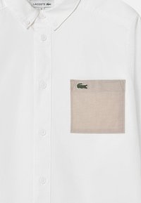 Camicia bianca con bottoni e tasca sul petto beige, caratterizzata da un logo di coccodrillo ricamato in verde. Tessuto liscio, design del colletto classico.