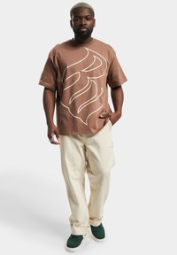 Maglietta di cotone marrone con design astratto bianco, abbinata a pantaloni con coulisse crema chiaro e sneakers verde scuro.