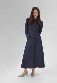 Giacca in denim scuro con chiusura a bottoni e maniche a sbuffo abbinata a una lunga gonna in denim. L'outfit è completato da tacchi grigi.