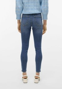 Jeans skinny en denim bleu foncé, avec une taille haute, deux poches arrière et une finition légèrement délavée. Portés avec des talons compensés crème.