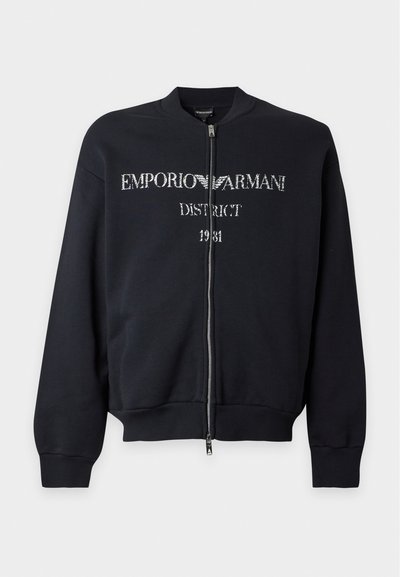 Blouson bomber noir zippé avec poignets et ourlet côtelés, avec texte et logo blanc délavé Emporio Armani District 1981 sur le devant.