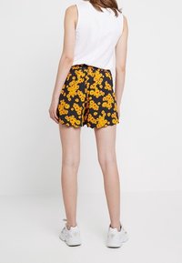 Shorts noirs avec un motif floral jaune, taille élastique et ourlet en dents de scie. Portés avec un top sans manches blanc et des baskets blanches.