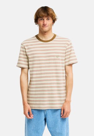 Jonge man met lichtbruin haar, gekleed in een beige-oranje gestreept T-shirt met een bruine kraag en lichtblauwe spijkerbroek, staand tegen een witte achtergrond.