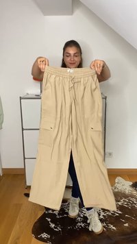 Kvinde holder beige cargobukser med lommer i et rum med hvide vægge, trægulv og et ko-skinds tæppe.