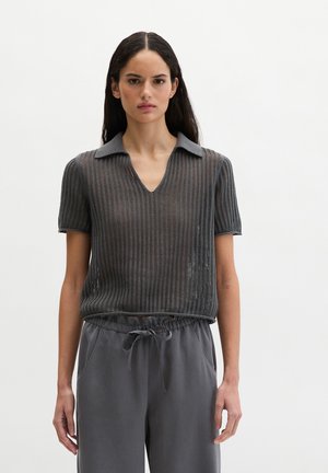 Vrouw met lang donker haar, gekleed in een grijs, opengebreid topje met korte mouwen en een kraag, en grijze broek met trekkoord, staand tegen een effen achtergrond.