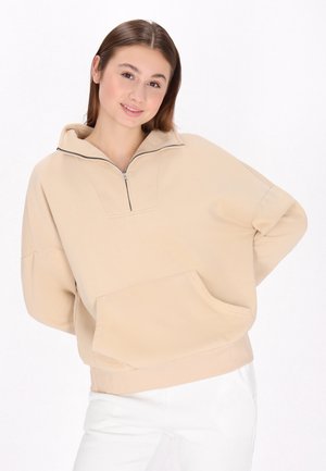 Beige oversized sweatshirt med en halv-zip krave, stor frontlomme, ribbede manchetter og blød tekstur. Bæres med hvide bukser.