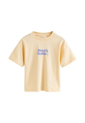 Copil tânăr blond purtând un tricou galben "Mama's Bestie" și blugi albaștri, întinzând mâna înainte cu o expresie concentrată.