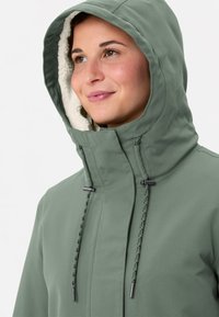 Donna che indossa una giacca invernale verde salvia con cappuccio, foderata in bianco di pile, che guarda leggermente in alto con un sorriso gentile.