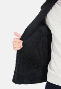 Giacca in pile nera con chiusura a zip, che presenta un interno ed esterno morbidi e testurizzati. Mano visibile che tiene la giacca aperta.