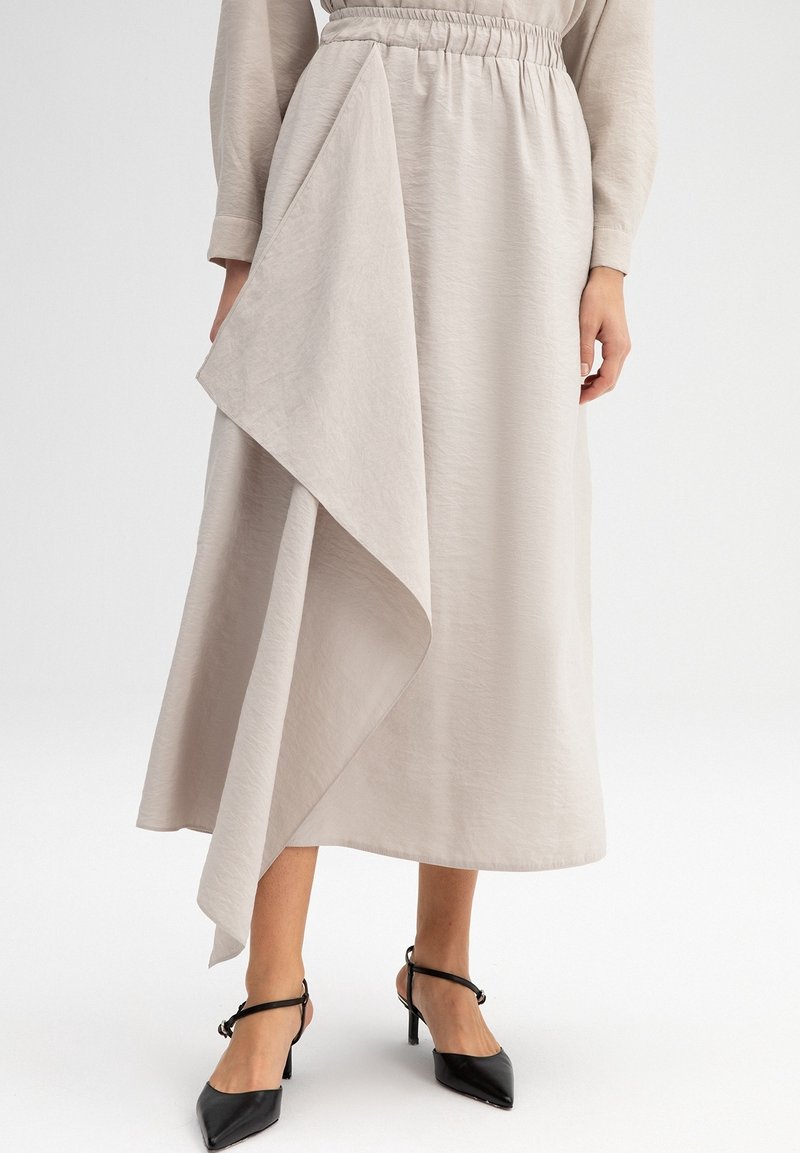 Jupe midi beige avec taille élastique et panneau drapé asymétrique, portée avec des talons noirs à bride cheville et bout pointu sur fond blanc.