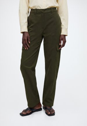 Personne portant un pantalon cargo vert olive, un t-shirt à manches longues jaune rayé et des sandales noires à bout carré sur un fond uni.