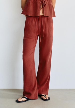 Nő laza rozsdaszínű, texturált nadrágot visel, melynek derékpántja zsinóros, fekete papucsokkal áll egy egyszerű fal előtt.