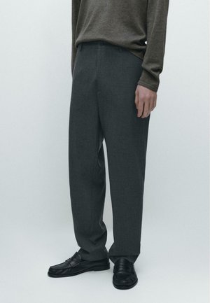 Uomo che indossa pantaloni tailored grigio scuro, maglia marrone a maniche lunghe e mocassini neri in pelle su uno sfondo chiaro uniforme.