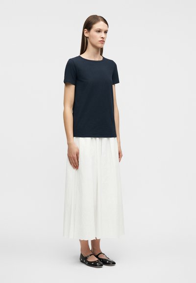 Γυναίκα που στέκεται φορώντας μπλε σκούρο πουκάμισο με κοντά μανίκια, λευκή πλισέ midi φούστα και μαύρα παπούτσια Mary Jane με μεταλλικές τρύπες.